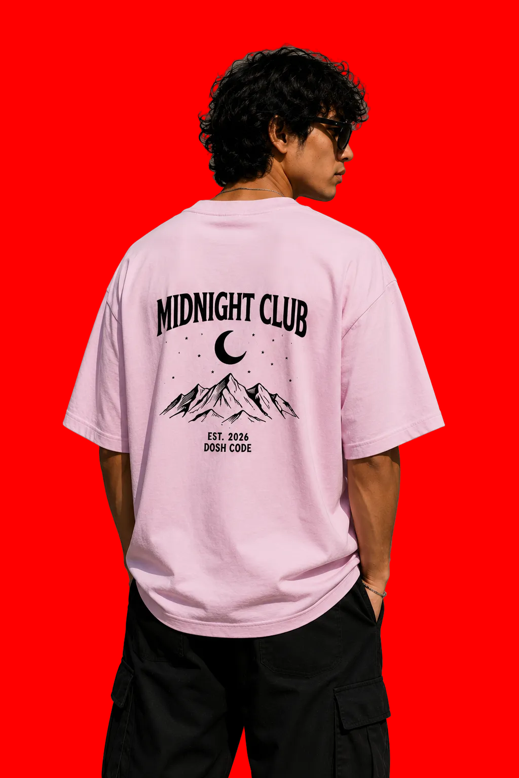 A man wearing a light pink graphic t-shirt with a black 'MIDNIGHT CLUB' mountain and moon design on the back, paired with black cargo pants, standing against a concrete wall.