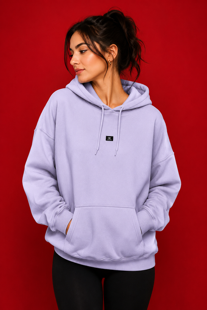 A woman wearing a lavender purple pullover hoodie with a small black square logo patch on the chest, posed against a solid red studio background.
